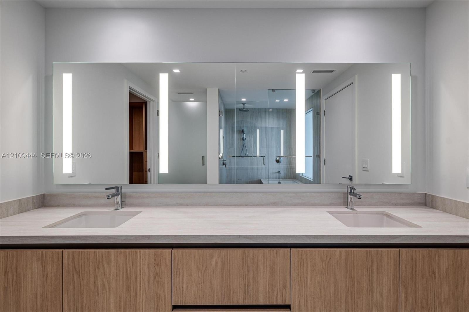 Dual-vanity cabinet with wood-finish cabinetry, neutral stone-composite countertop, integrated dual undermount sinks, and full-width mirror featuring 