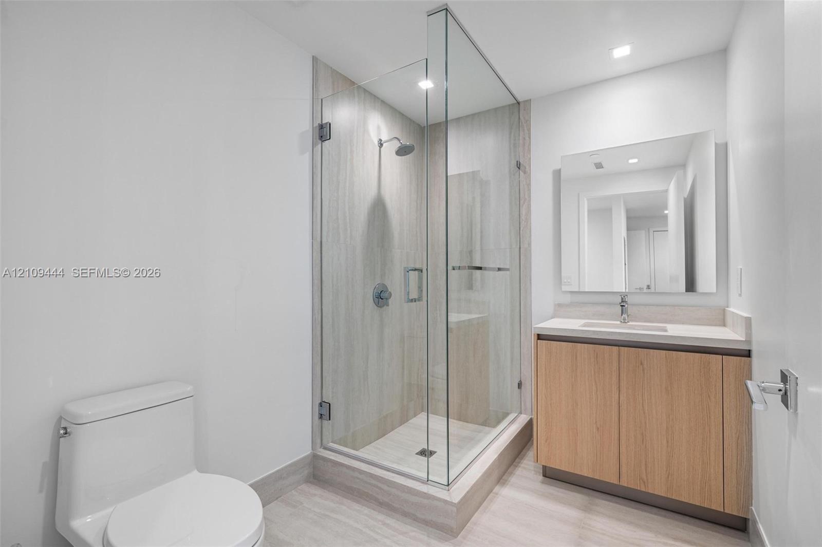 Walk-in glass shower with floor-to-ceiling tile, light wood-finish cabinetry, rectangular vessel sink, surface-mounted vanity mirror, and recessed lig