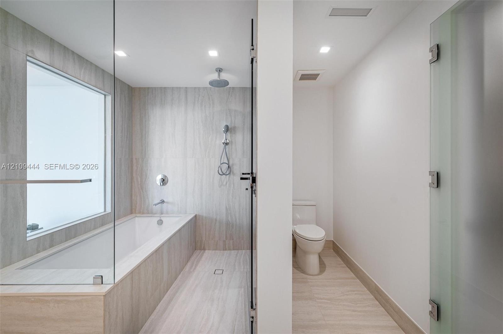 Bathroom featuring floor-to-ceiling stone-finish tile, glass-enclosed soaking tub with rain-style shower head, recessed lighting, and a private water 