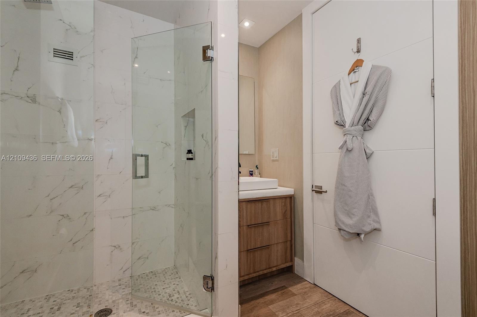 Walk-in shower featuring glass paneling, marble-style tile walls, and a pebble-stone floor, paired with a wood-finish vanity, vessel sink, and solid-c