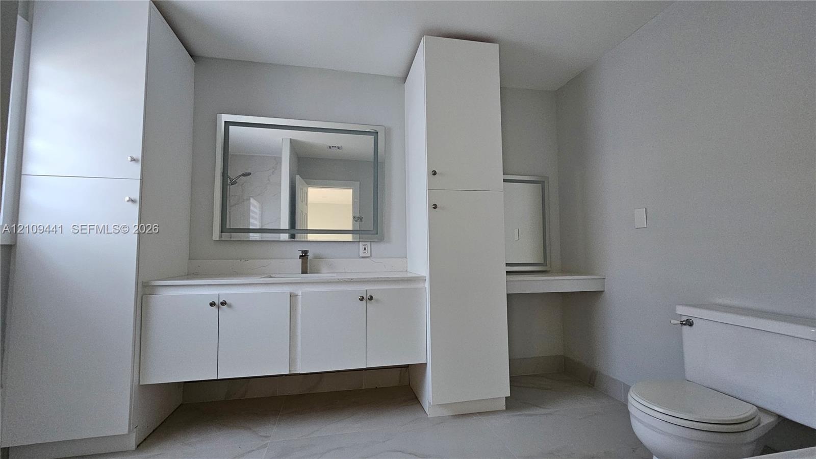 Bathroom featuring white cabinetry with integrated floor-to-ceiling storage, stone-surface countertop, rectangular wall mirror with integrated lightin