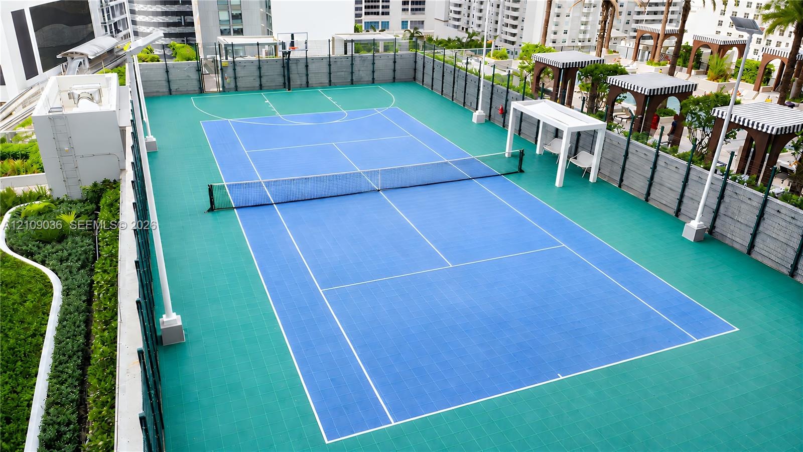 Rooftop multi-use athletic court featuring a synthetic surface, integrated basketball hoop, tennis net, and surrounding perimeter fencing with shade s