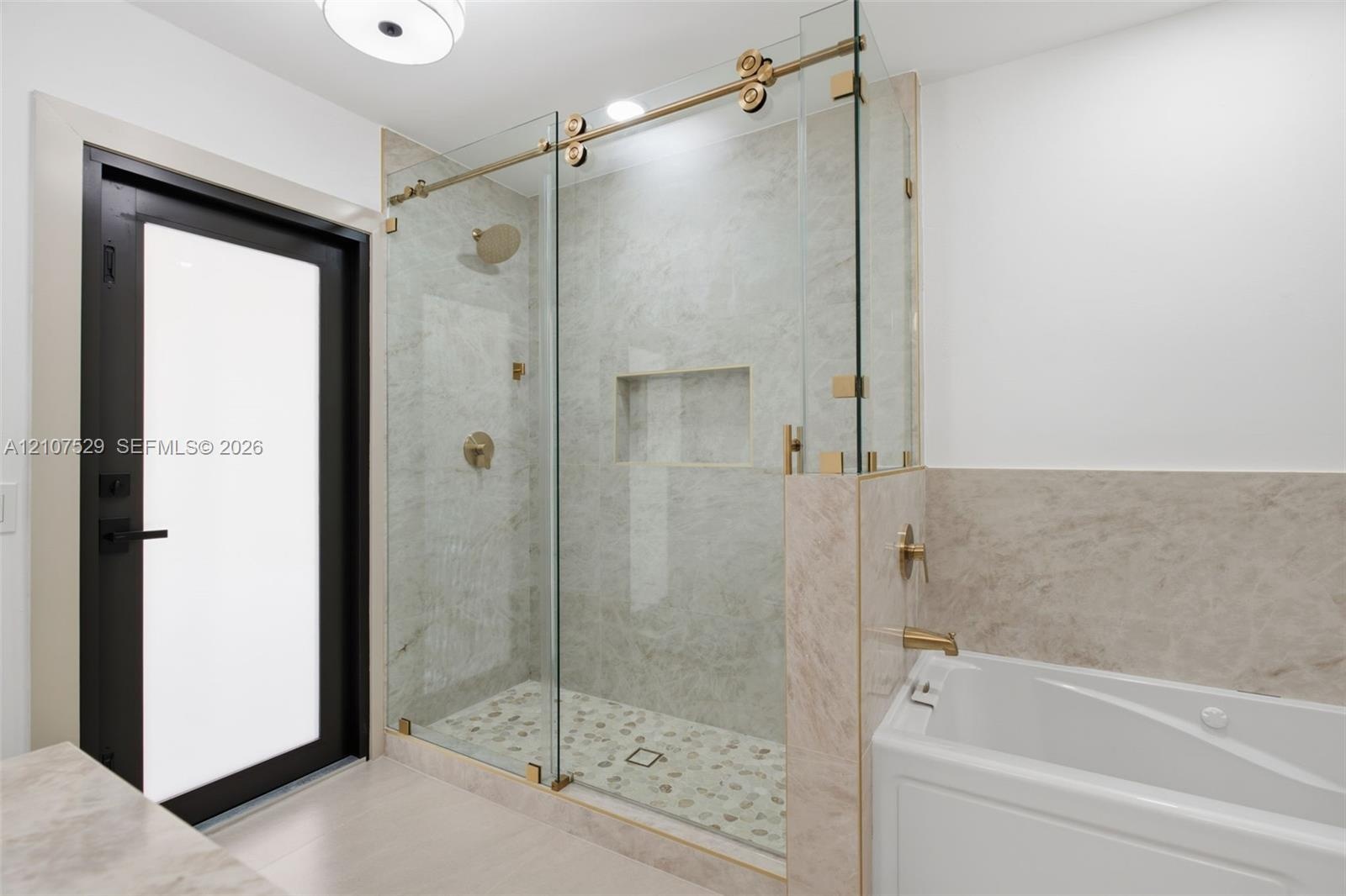 Walk-in shower with frameless glass enclosure, gold-toned hardware, stone-tile surround, and recessed wall niche, featuring a separate white soaking t