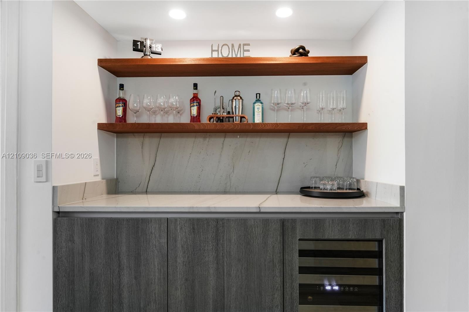 Custom wet bar featuring charcoal-toned cabinetry, a stone-patterned countertop and backsplash, dual wood-finish floating shelves, and a built-in wine