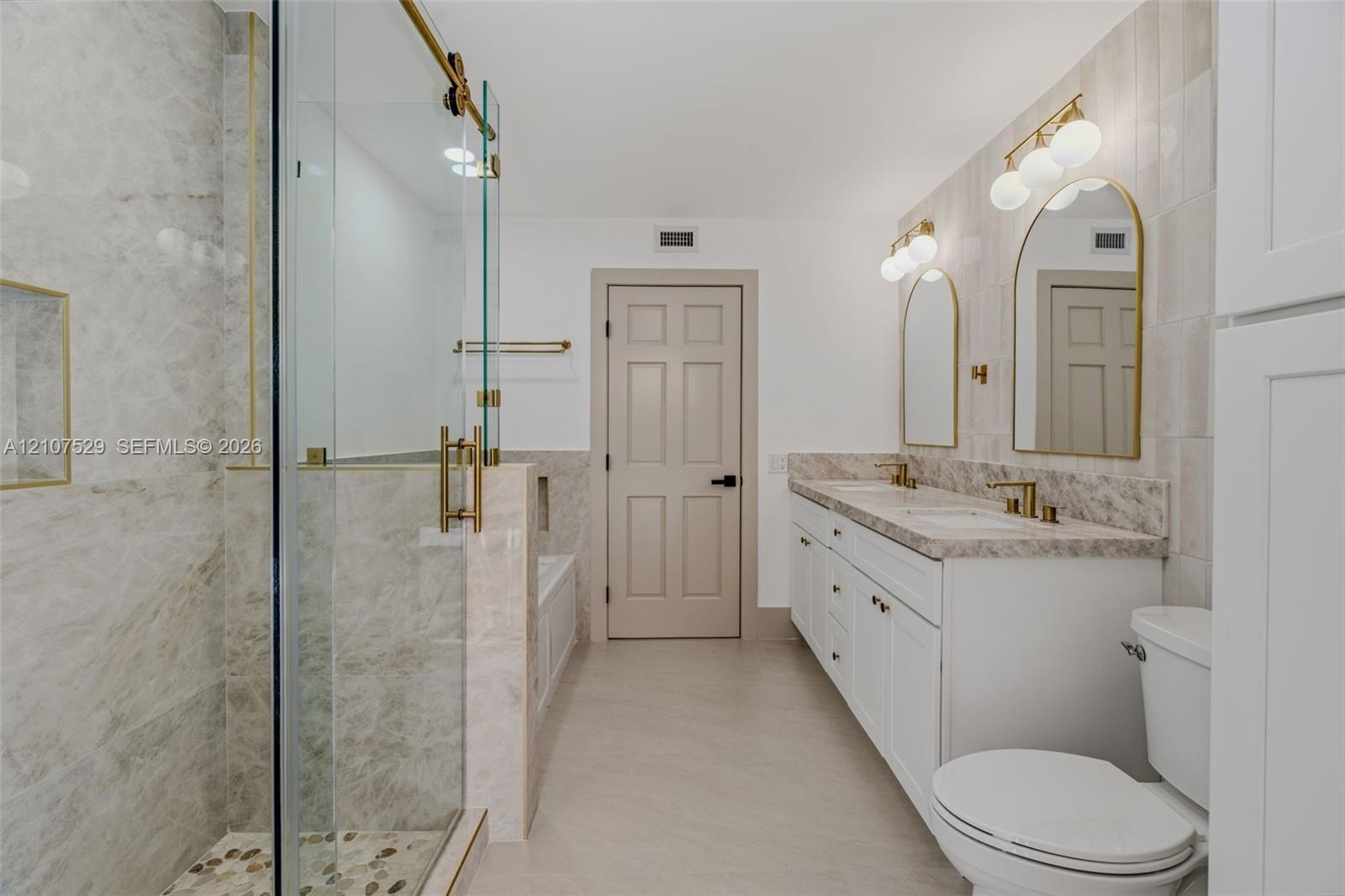 Double vanity with a stone countertop, arched mirrors, brass-finish fixtures, and floor-to-ceiling ceramic tile wall detailing