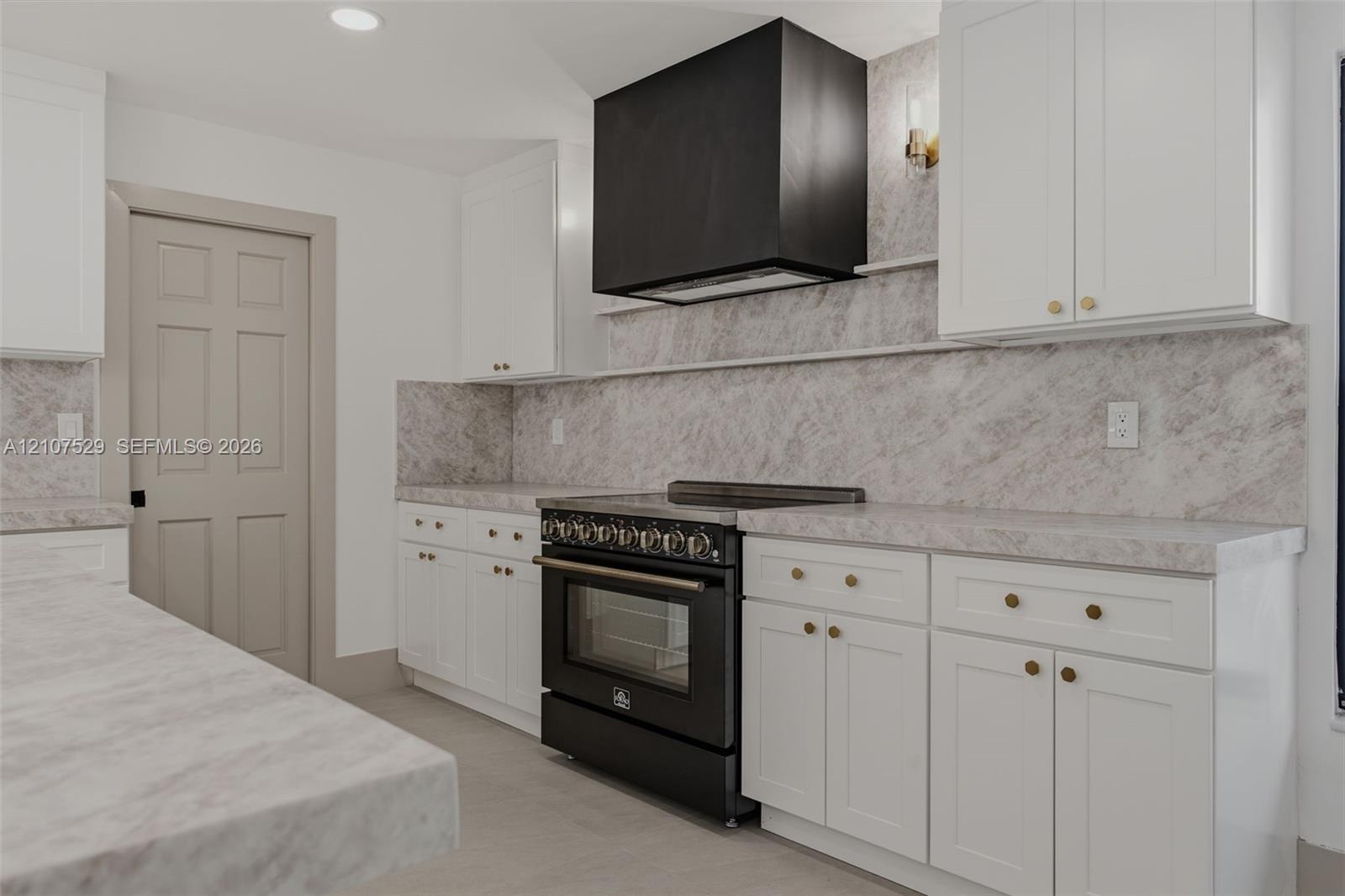Kitchen featuring white shaker cabinetry, stone-finish countertops and backsplash, black range with matching vent hood, and natural wood floating shel
