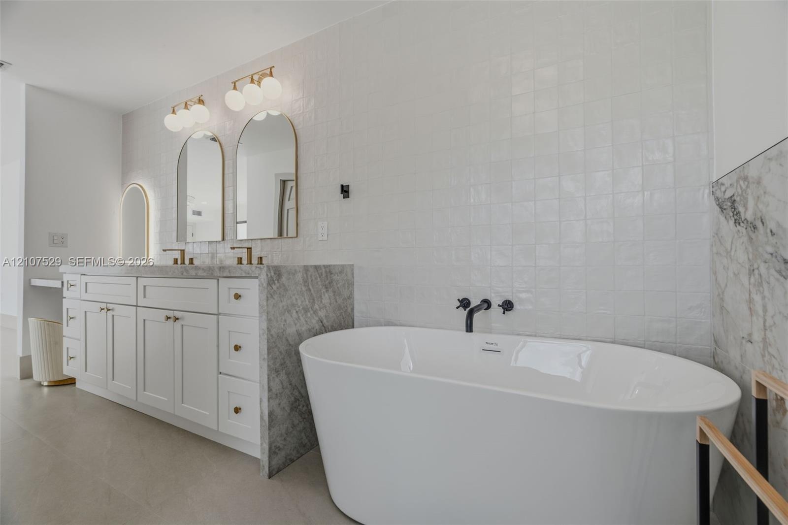 Dual-sink vanity with stone countertops, arched mirrors with integrated lighting, white cabinetry with brass hardware, free-standing soaking tub, and 