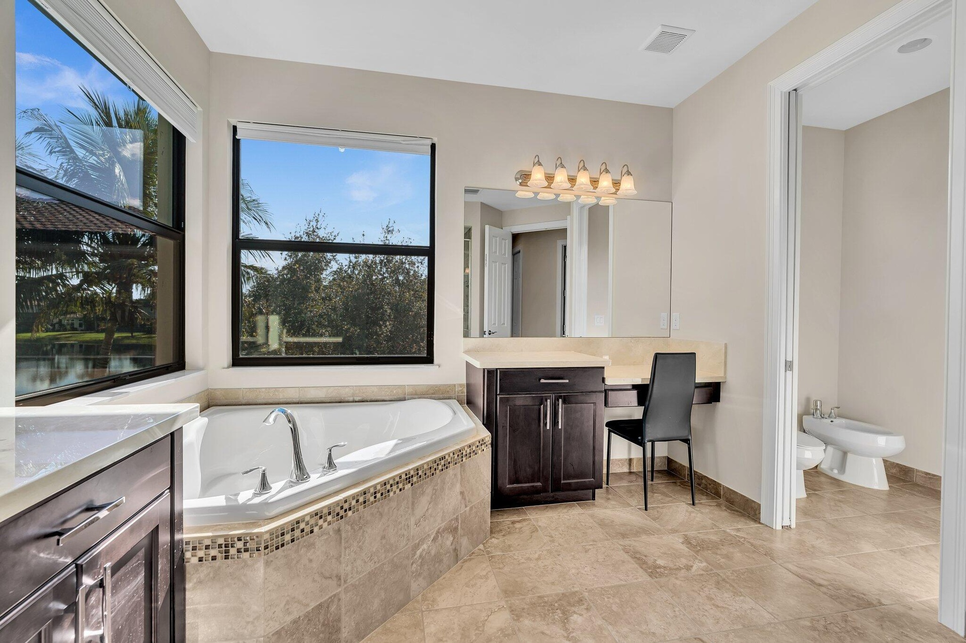 Corner soaking tub with mosaic tile surround, large windows, dark wood cabinetry with light countertops, and neutral tile flooring