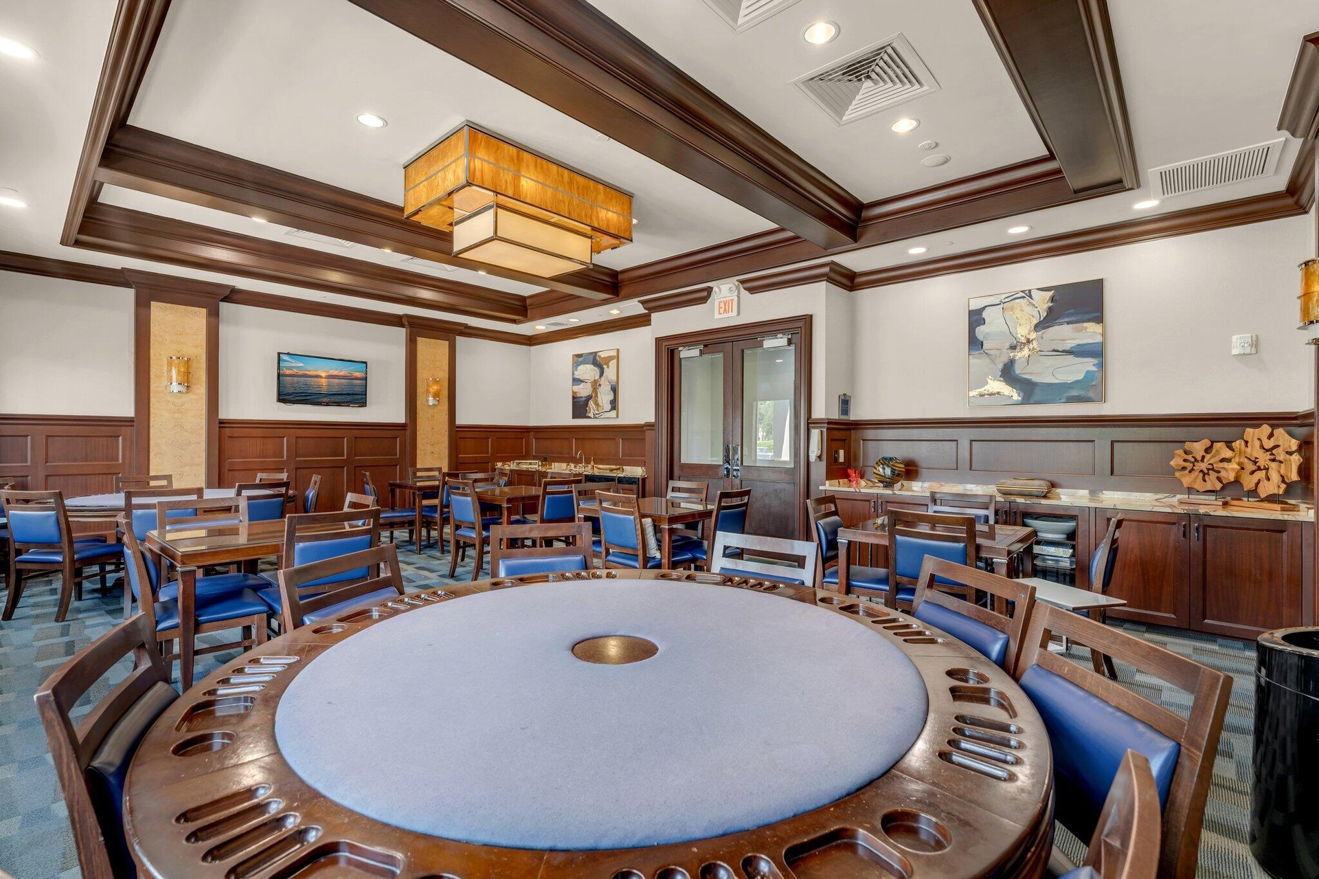 Spacious common area featuring a large round gaming table, multiple dining tables, wood-finish wall paneling, and built-in cabinetry with countertops