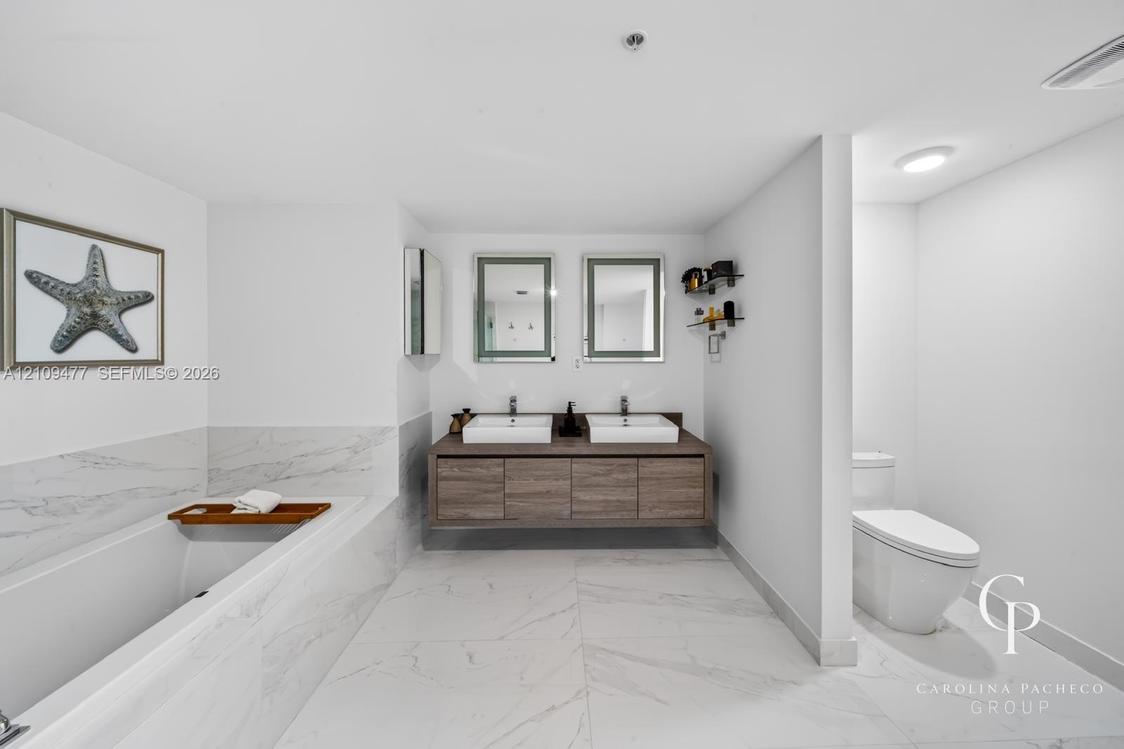 Floating dual-sink vanity with wood-finish cabinetry, rectangular vessel sinks, wall-mounted rectangular mirrors, soaking tub, and large-format tile f