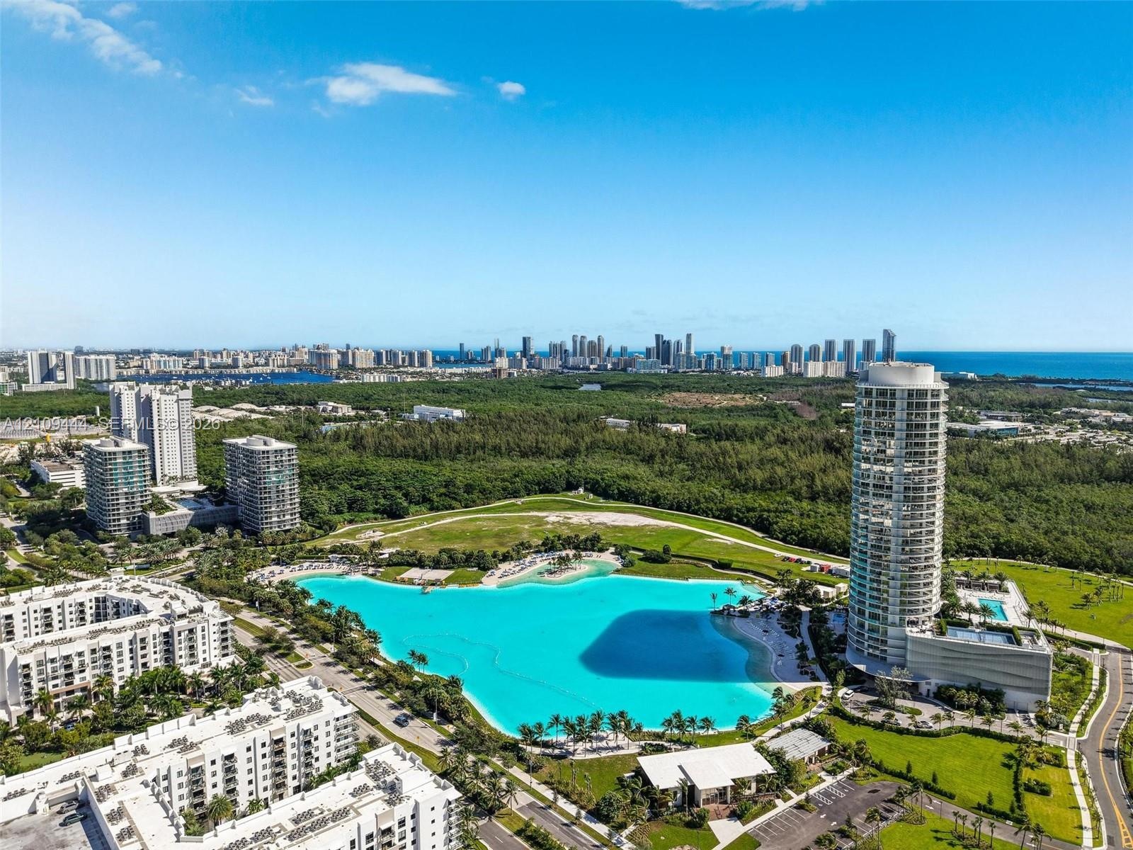 Aerial perspective of a high-rise residential tower featuring a large man-made lagoon with a sandy shoreline, surrounding parkland, and coastal city s
