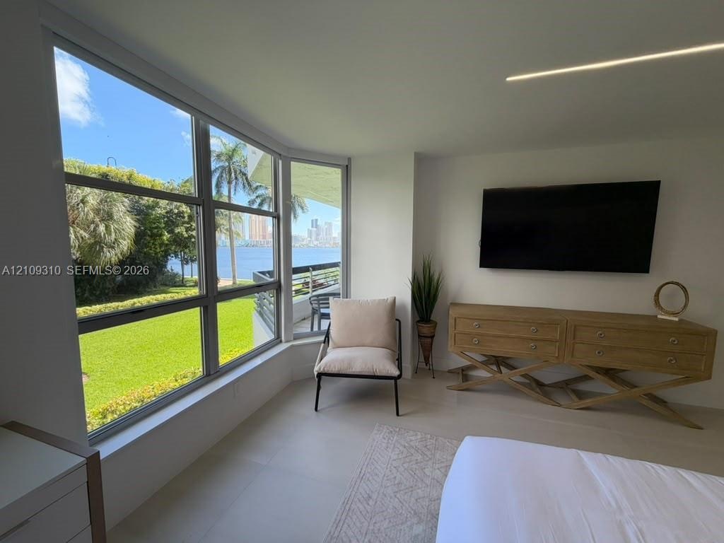 Floor-to-ceiling windows featuring waterfront scenery, neutral tile flooring, recessed linear lighting, and wall-mounted television console