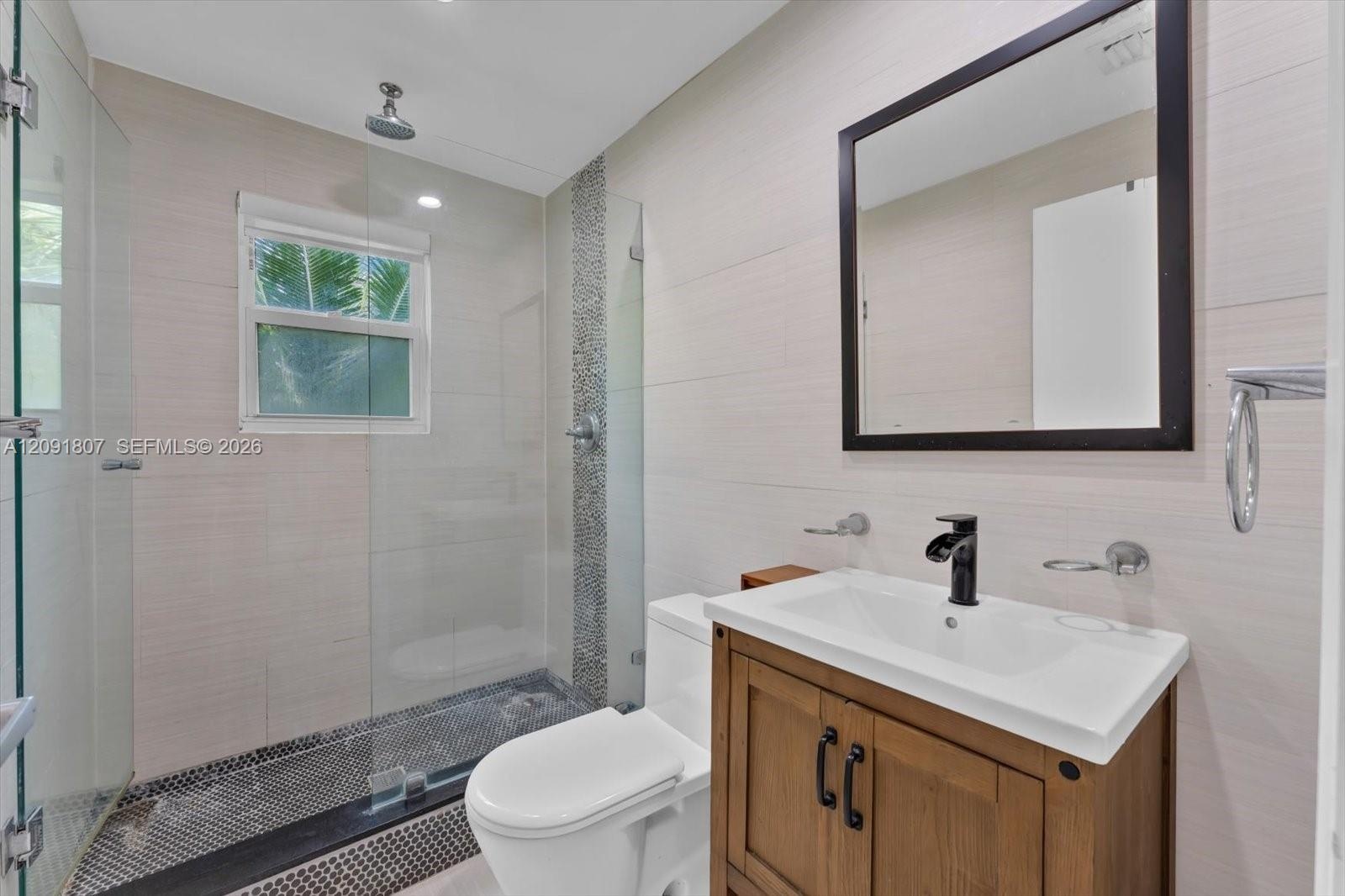 Bathroom featuring a wood-finish vanity with an integrated sink, matte black faucet, walk-in shower with a glass enclosure, pebble-tile flooring, and 
