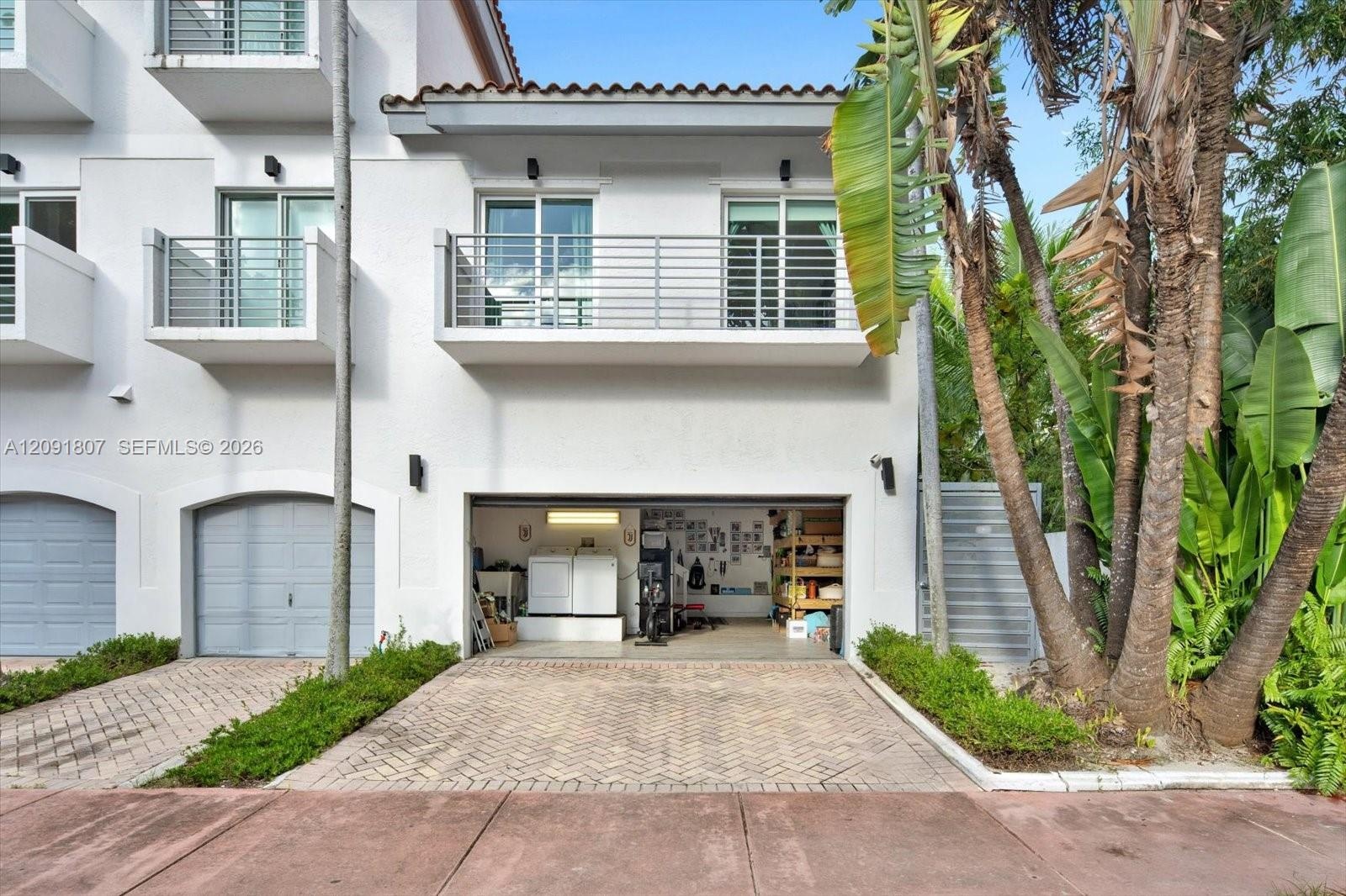 Stucco exterior featuring dual-level metal balconies, terracotta-style roof tile accents, a private herringbone-patterned paver driveway, and an attac