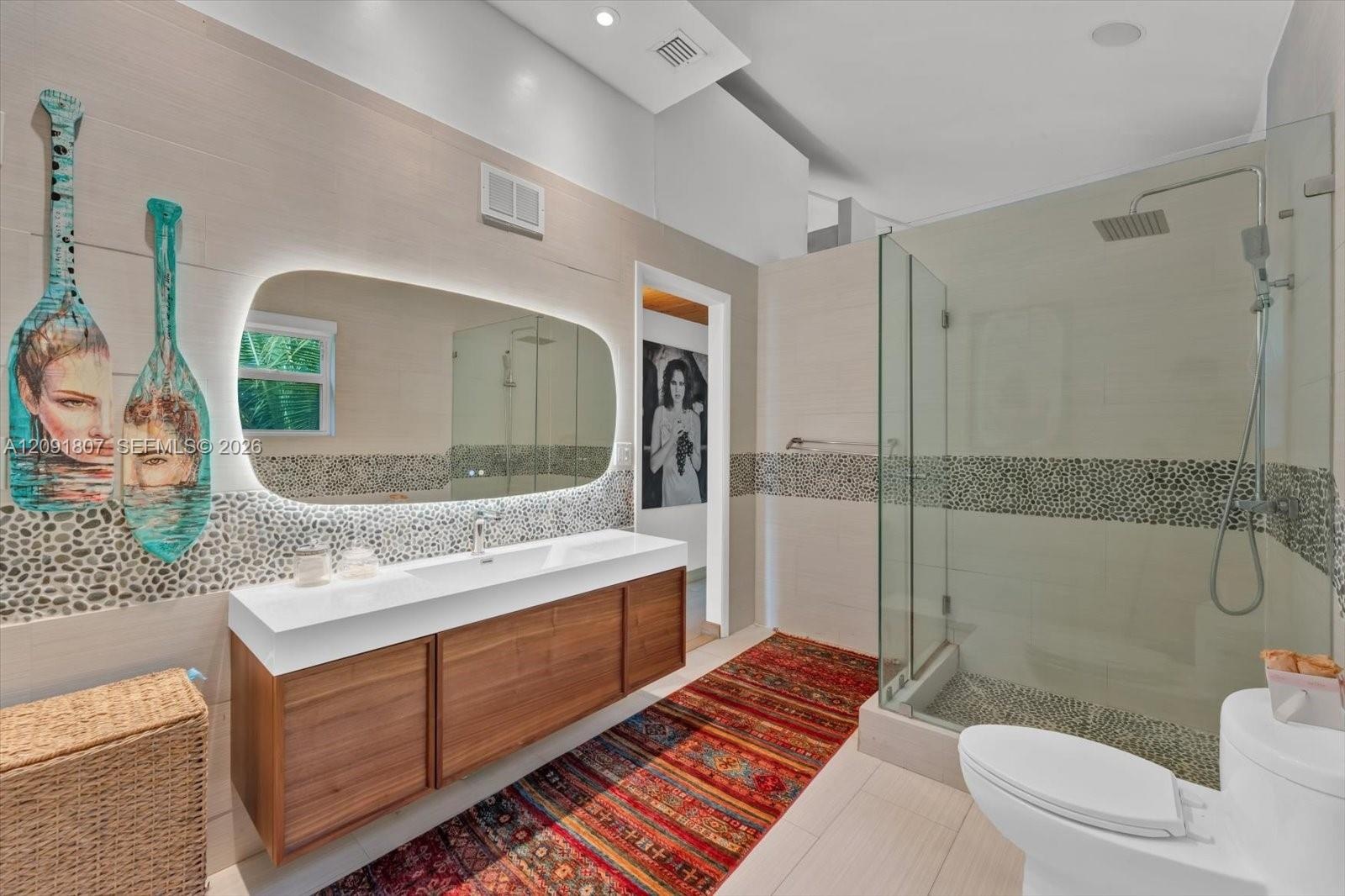 Floating wood-finish vanity with a white countertop and integrated sink, illuminated irregular-shaped vanity mirror, glass-enclosed shower with pebble