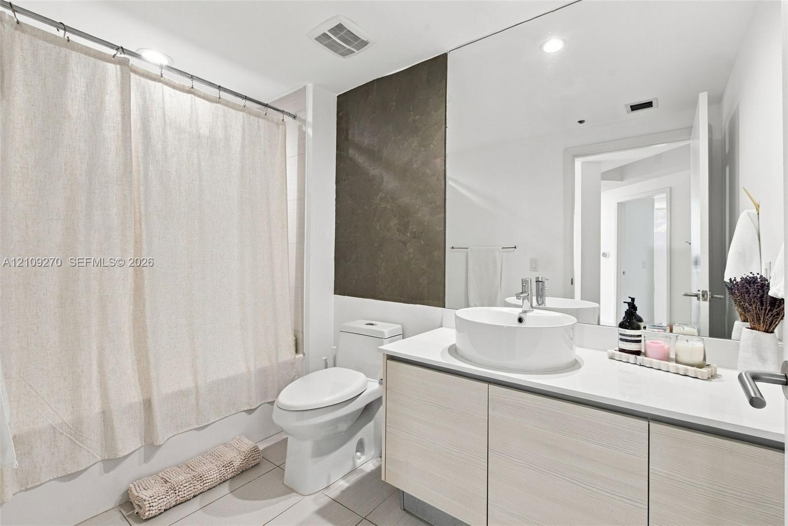 Bathroom featuring a vanity with wood-finish cabinetry, a white countertop, and a vessel sink, accompanied by a full-wall mirror, tiled flooring, and 