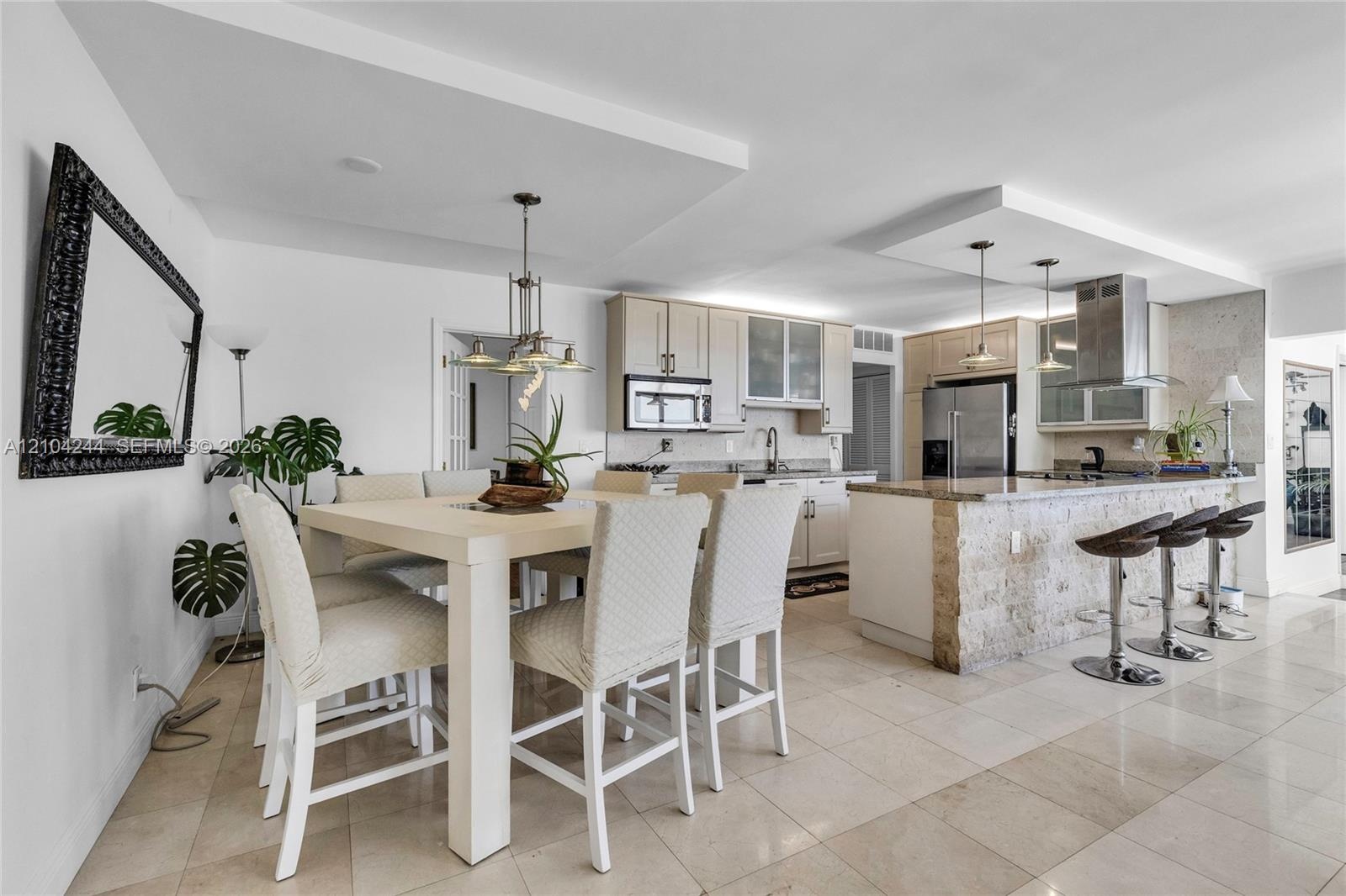 Open-concept kitchen featuring light wood cabinetry, stone-clad kitchen island, stainless steel appliances, tiered tray ceilings, and neutral tile flo