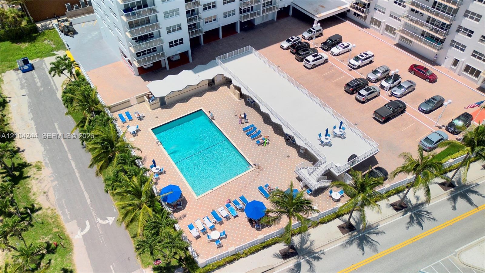Aerial view of the building exterior featuring an in-ground swimming pool, paved pool deck, multi-level parking area, and landscaping with palm trees