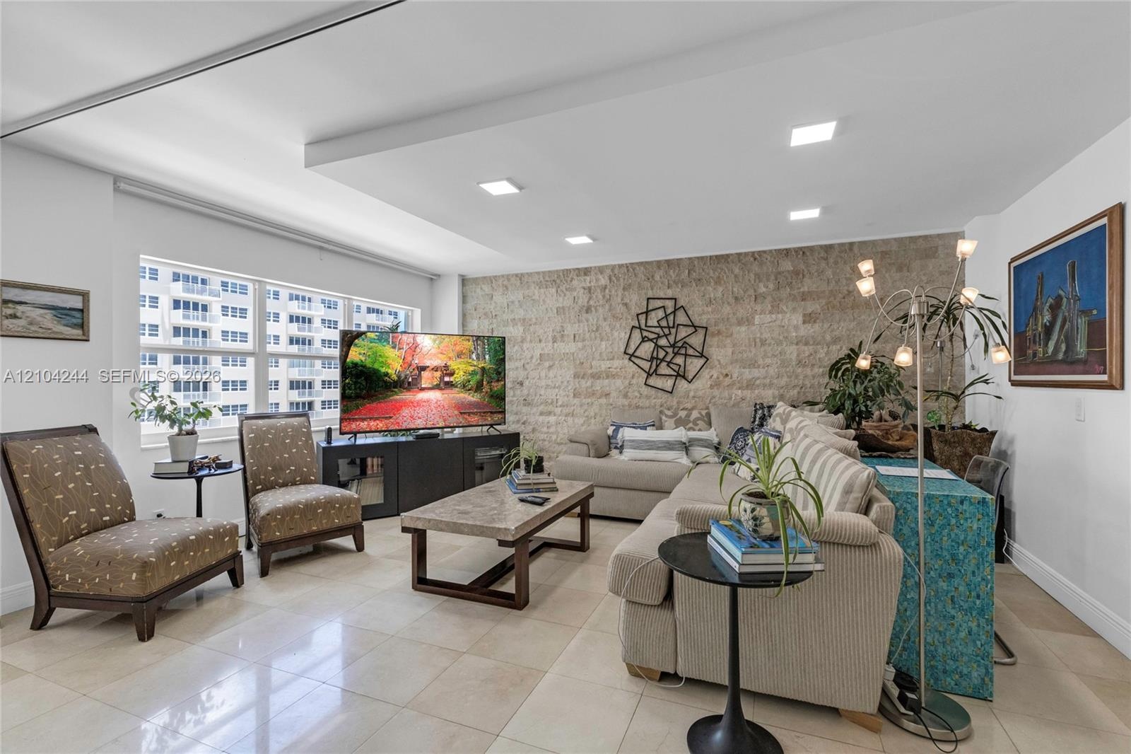 Living area featuring a textured stone accent wall, large-format tile flooring, a recessed tray ceiling with integrated lighting, and expansive window