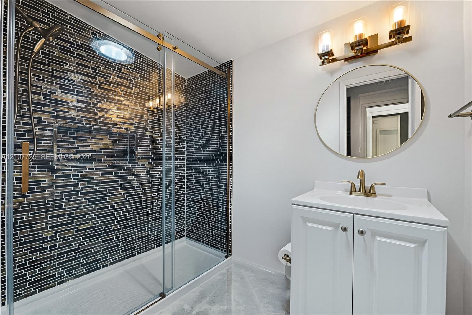 Bathroom featuring a walk-in shower with glass sliding doors and multi-tonal linear tile surround, brass-finish fixtures, white vanity cabinet with an
