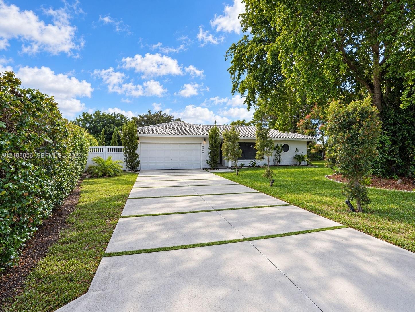 Single-story residence featuring a light-toned stucco exterior, a segmented concrete driveway, a multi-car garage, a tiled roof, and mature perimeter 