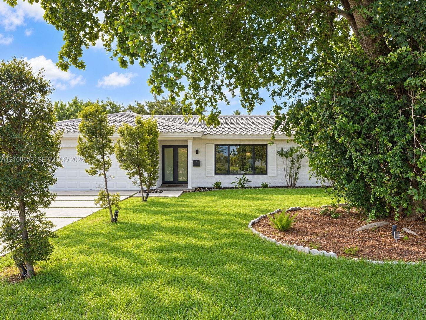 Single-story residence featuring a white painted brick exterior, tile roof, expansive front lawn, landscaped mulch garden beds with stone border, and 