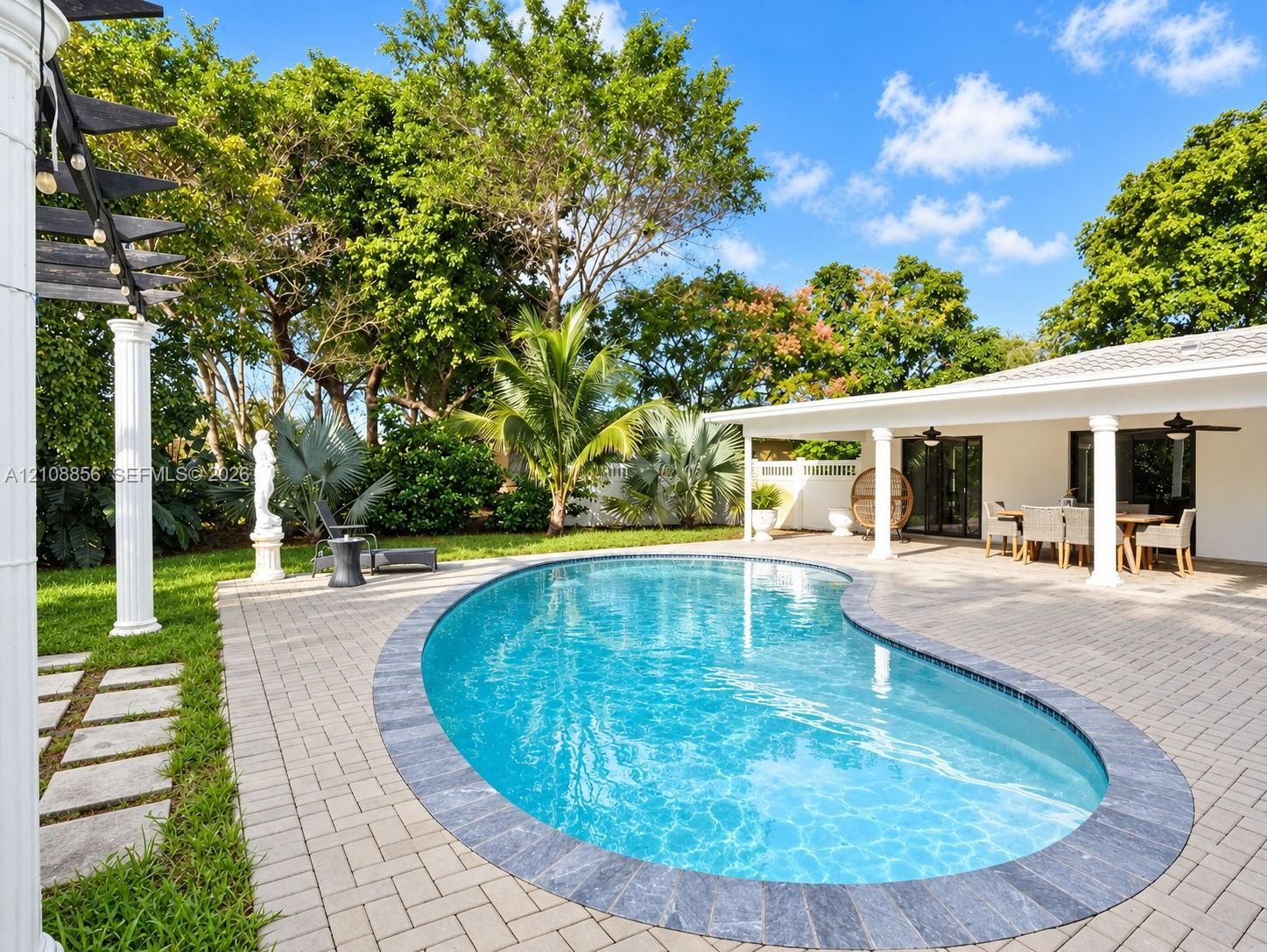 Kidney-shaped swimming pool with stone coping, expansive paver patio, covered porch with classical columns, and mature landscaping