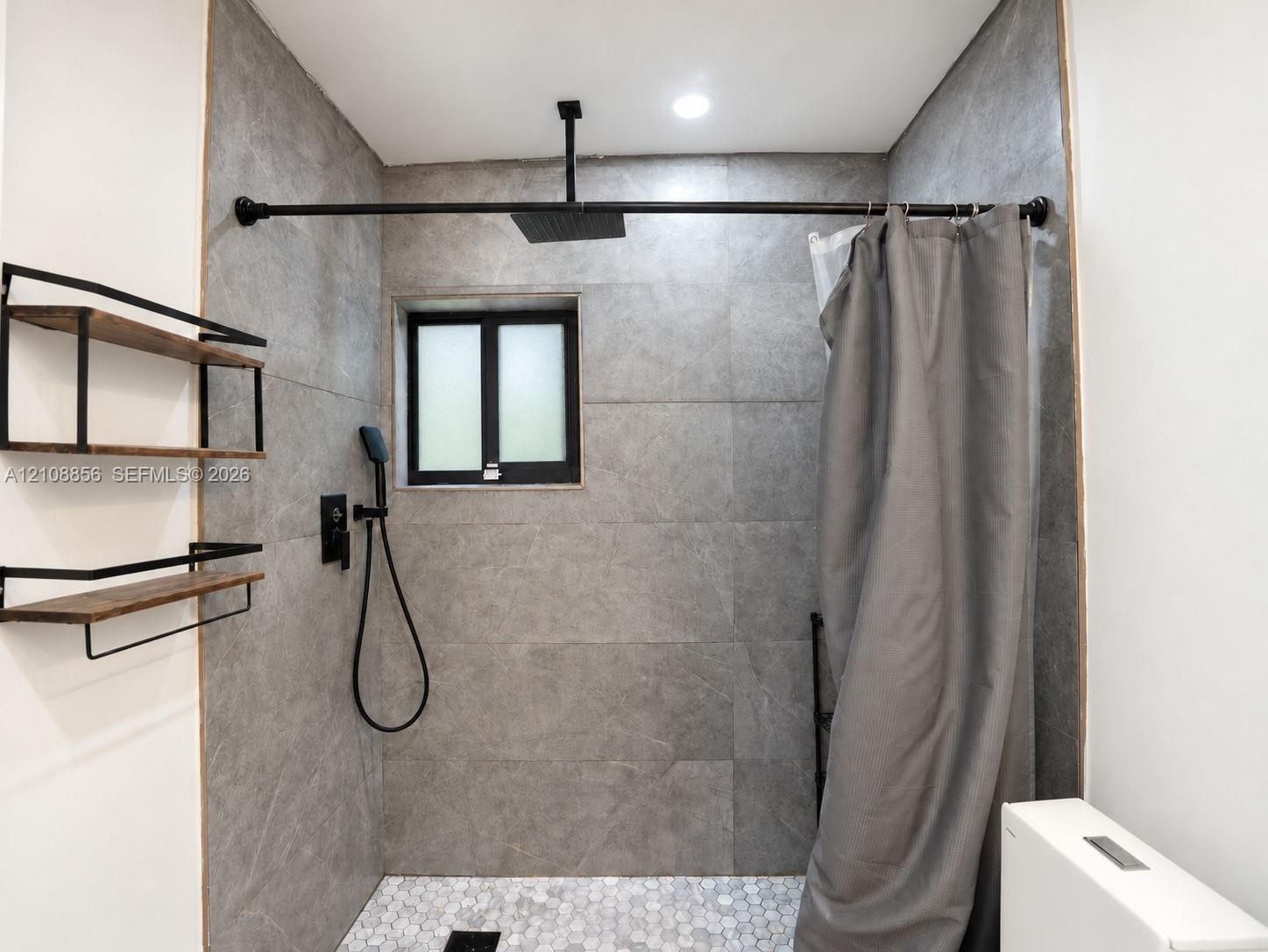 Shower area featuring large-format grey tile walls, a white hexagonal tile floor, matte black rainfall showerhead with handheld sprayer, and built-in 