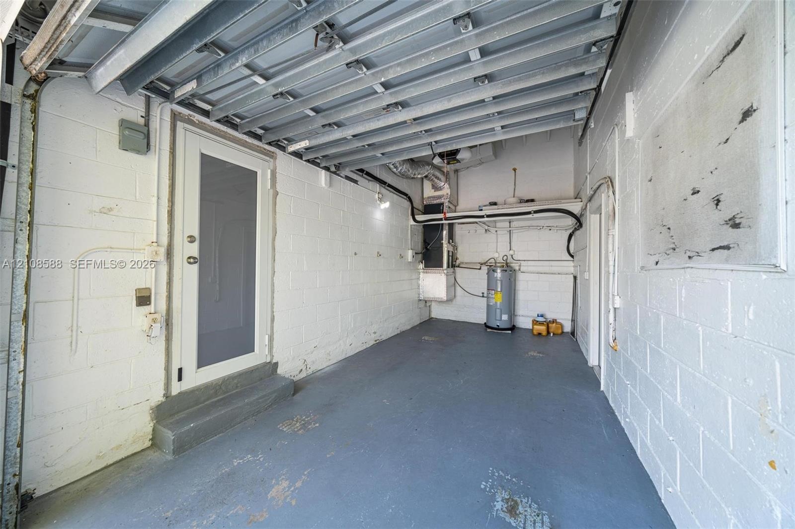 Enclosed garage featuring painted concrete block walls, painted concrete flooring, exposed ceiling joists, a dedicated utility area with a water heate