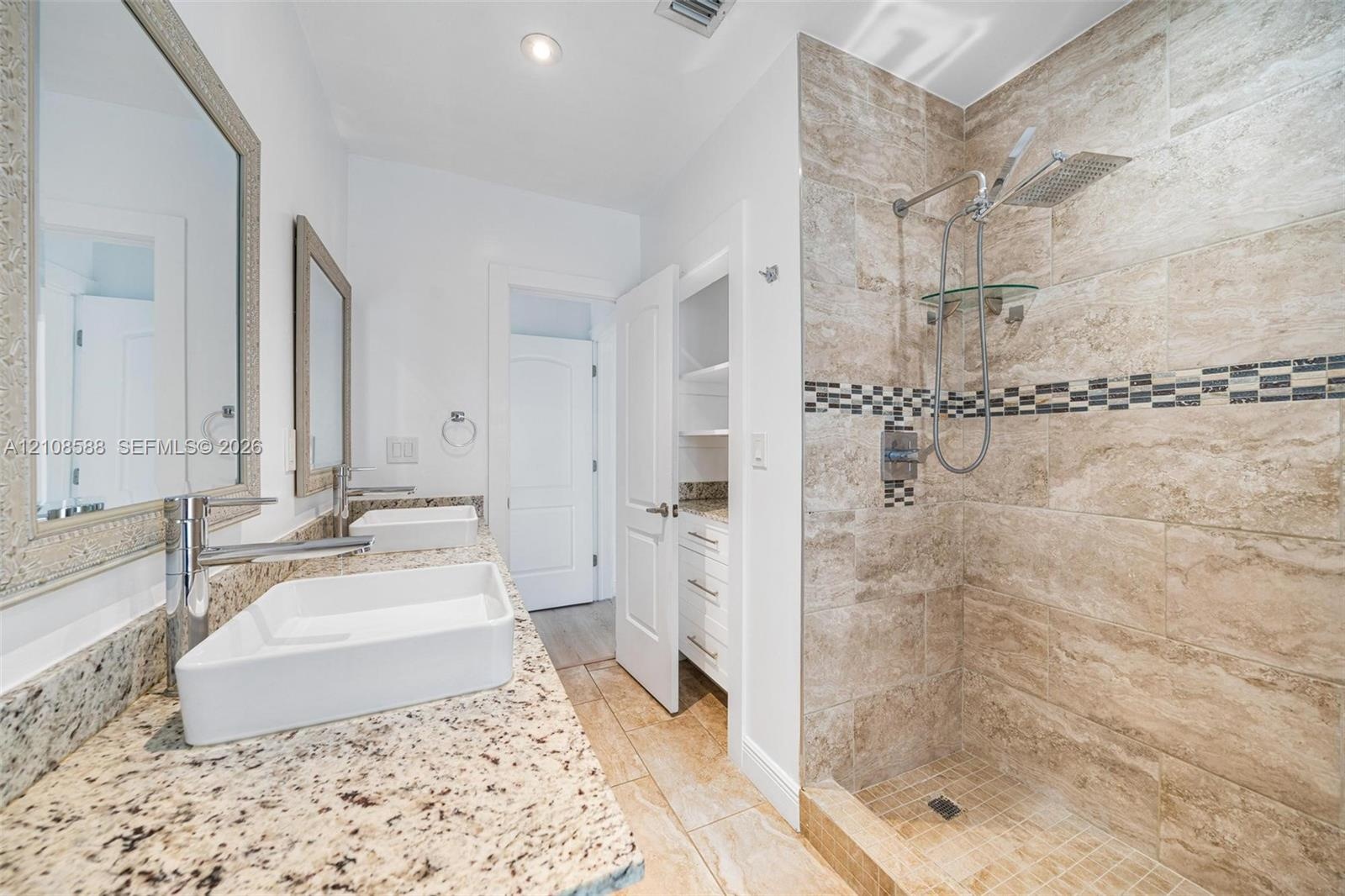 Dual vanity with rectangular vessel sinks and stone countertop, walk-in shower with natural stone tile and mosaic inlay, built-in linen cabinetry, and