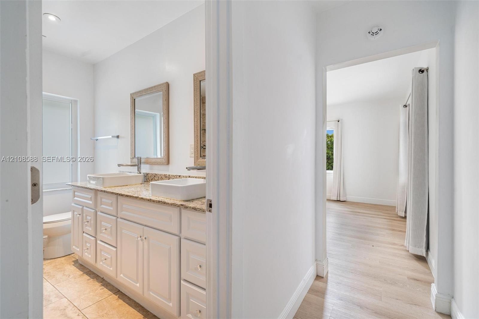 Dual-sink vanity with white cabinetry, stone-finish countertop, and rectangular vessel sinks, complemented by wood-finish flooring in the hallway