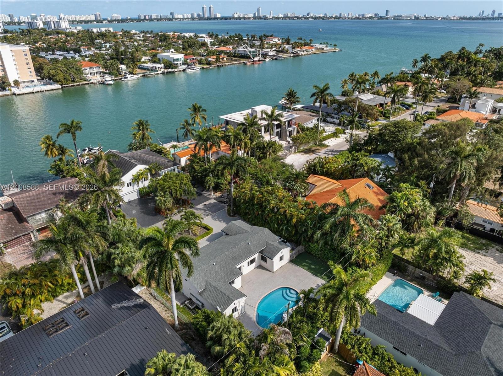 Aerial perspective featuring a single-story residence with a shingle roof, paved patio, and in-ground swimming pool situated on a waterfront lot with 