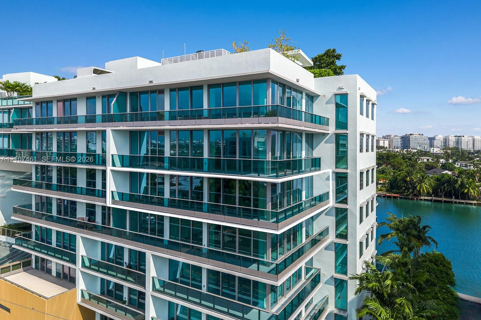 Modern multi-story building featuring white stucco facade, expansive floor-to-ceiling windows, wrap-around glass-railed balconies, and a rooftop terra