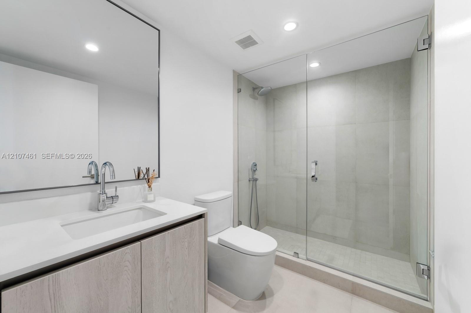 Bathroom featuring a wood-finish vanity with a stone countertop, an integrated rectangular sink, a wall-to-wall mirror, and a walk-in shower with a gl
