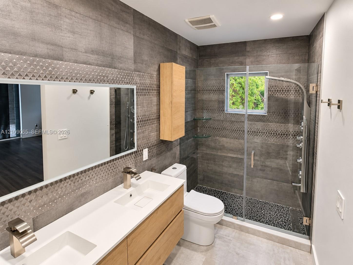 Bathroom featuring a dual-sink vanity with a white countertop, large-format grey wall tile with a geometric accent band, glass-enclosed shower with bo