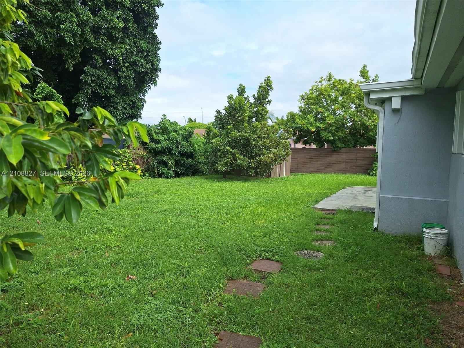 Backyard featuring a grass lawn, paved patio area, stone paver pathway, mature trees, and perimeter fencing