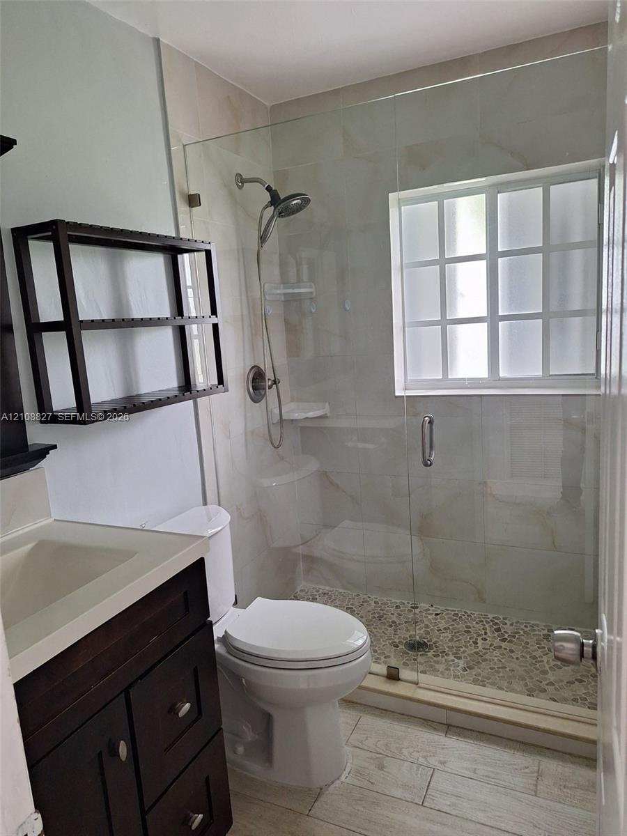 Bathroom featuring a glass-enclosed shower with a pebble stone floor, wood-finish flooring, a dark wood vanity with a solid-surface countertop, and a 