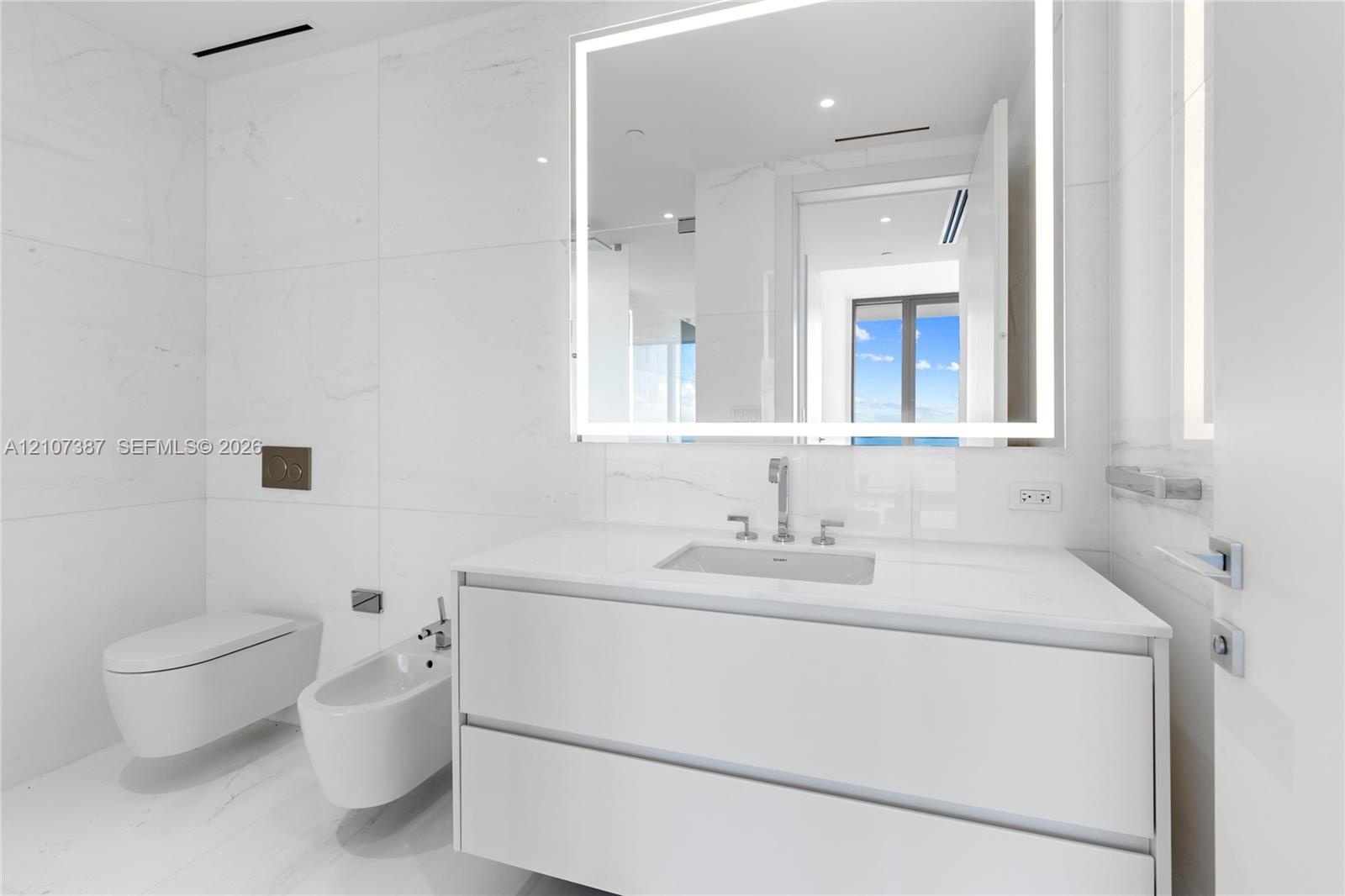 Modern floating vanity with a seamless countertop, wall-mounted commode and bidet, large-format tile wall cladding, and an integrated LED backlit mirr