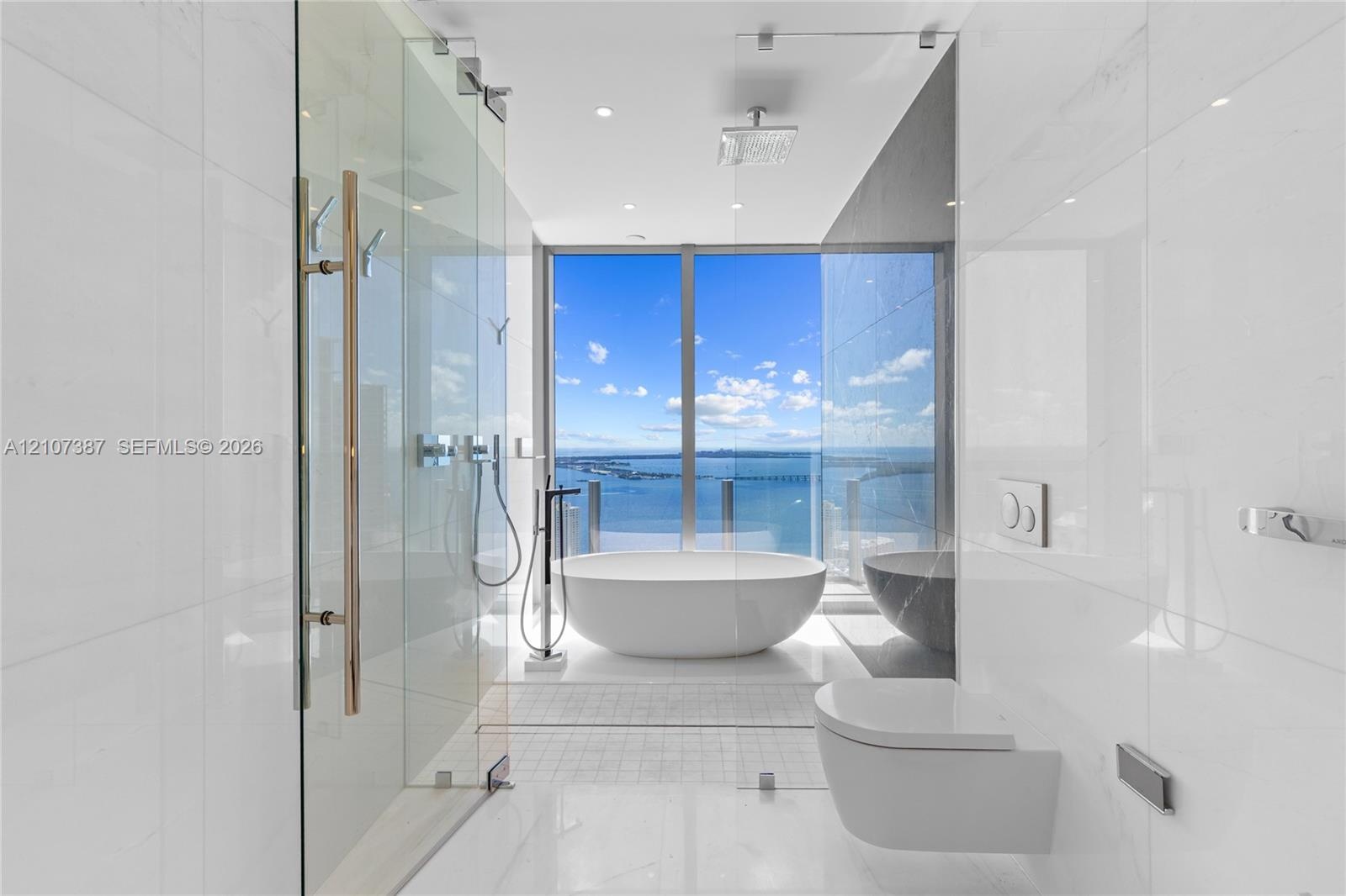Freestanding soaking tub, floor-to-ceiling glass-enclosed shower, wall-mounted commode, large-format tile flooring, and floor-to-ceiling windows with 
