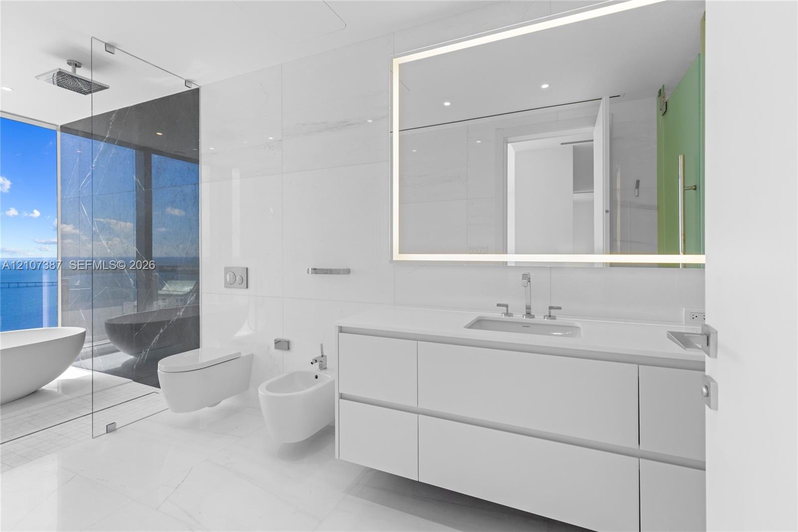 Modern floating vanity with a white countertop and rectangular sink, LED-backlit mirror, wall-mounted commode and bidet, and floor-to-ceiling glass-en