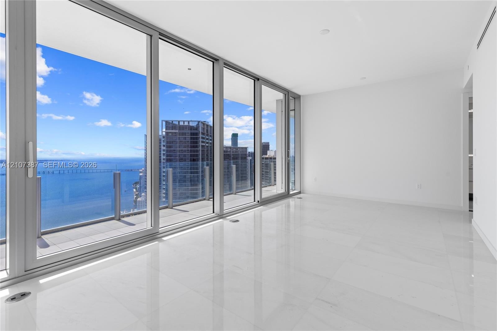 Floor-to-ceiling glass panel windows, wide-plank tile flooring, and a glass-railed exterior balcony featuring a water and cityscape backdrop
