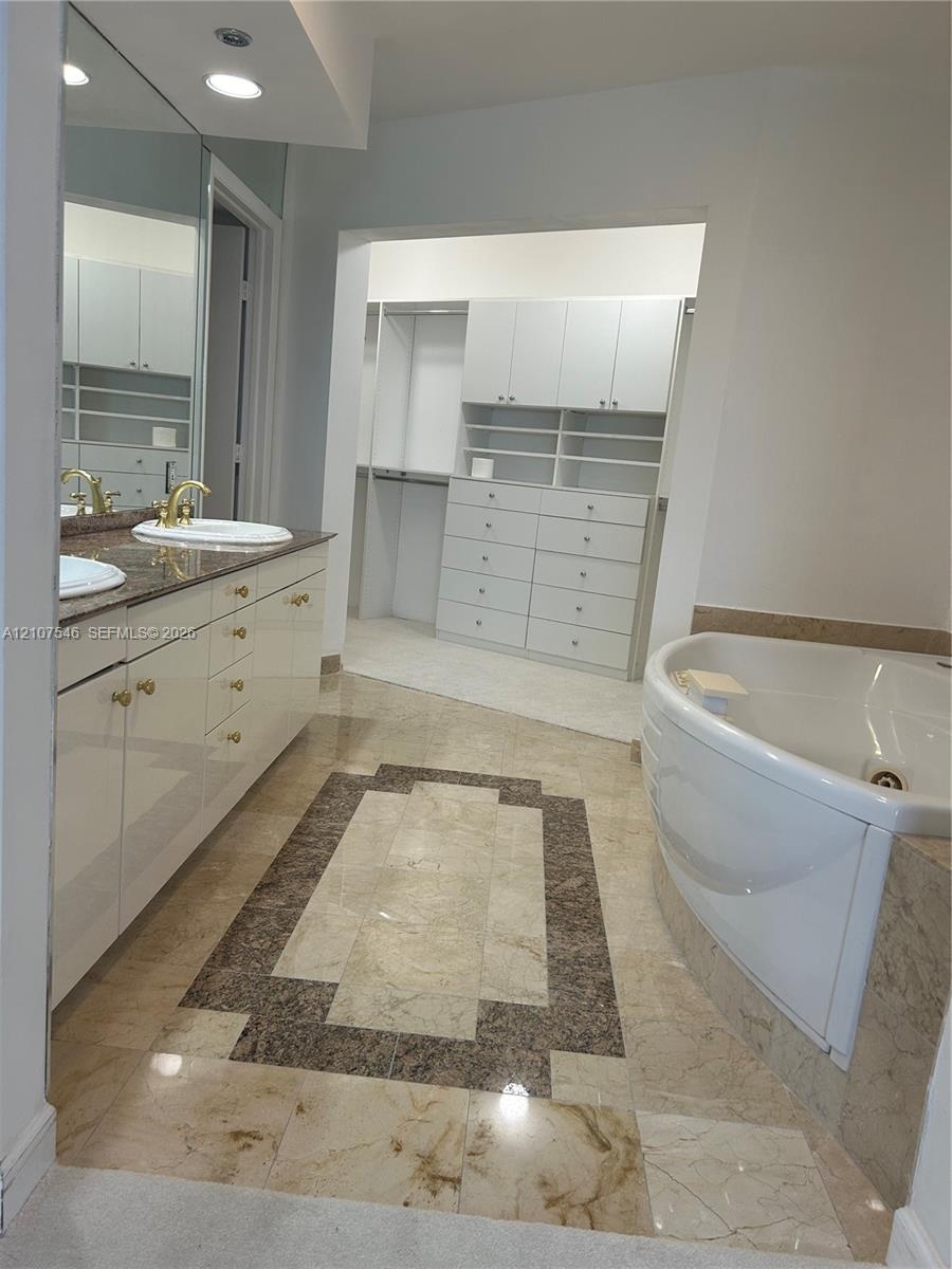 Dual-sink vanity with stone countertop and gold-tone fixtures, expansive wall mirror, jetted soaking tub with stone surround, and custom built-in ward