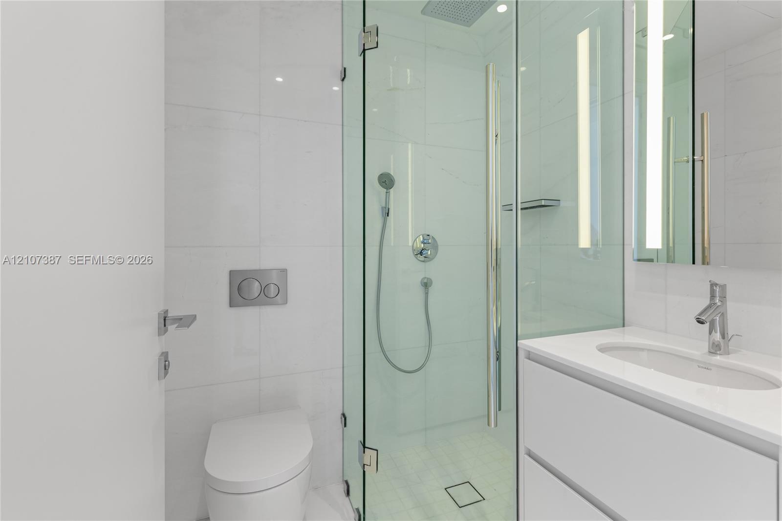 Bathroom featuring a glass-enclosed shower with a rainfall showerhead, wall-mounted dual-flush toilet, white vanity with an integrated sink, polished 