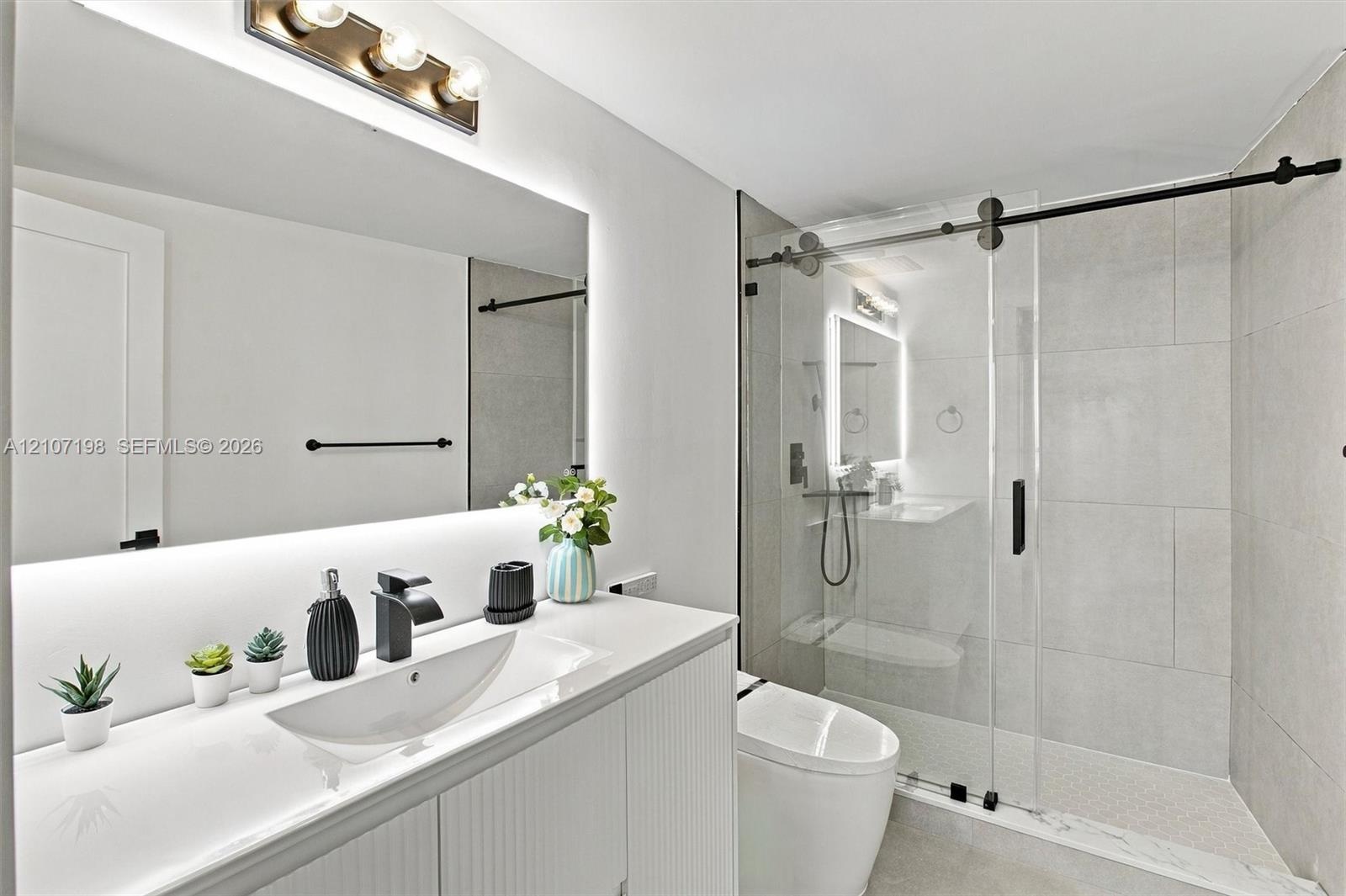 Bathroom featuring a white vanity with an integrated sink, matte black faucet, full-width mirror with integrated lighting, and a glass-enclosed shower