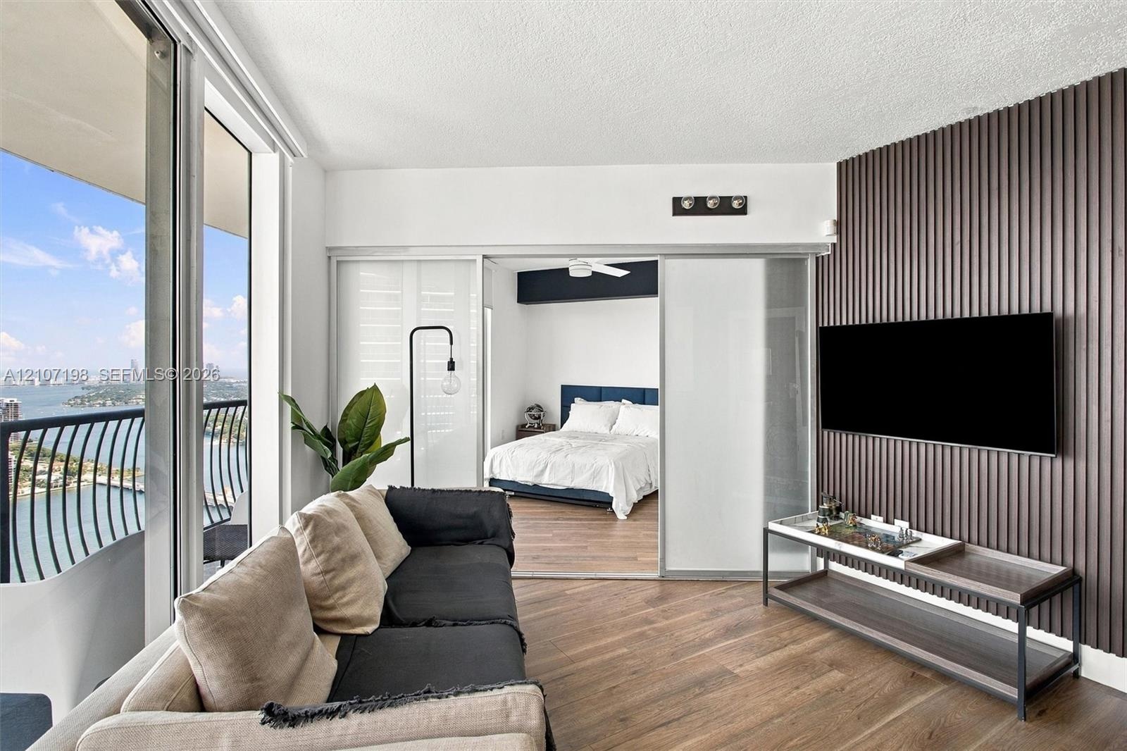 Floor-to-ceiling windows, wood-finish flooring, accent wall with vertical slat detailing, sliding glass room divider, and private balcony with water e
