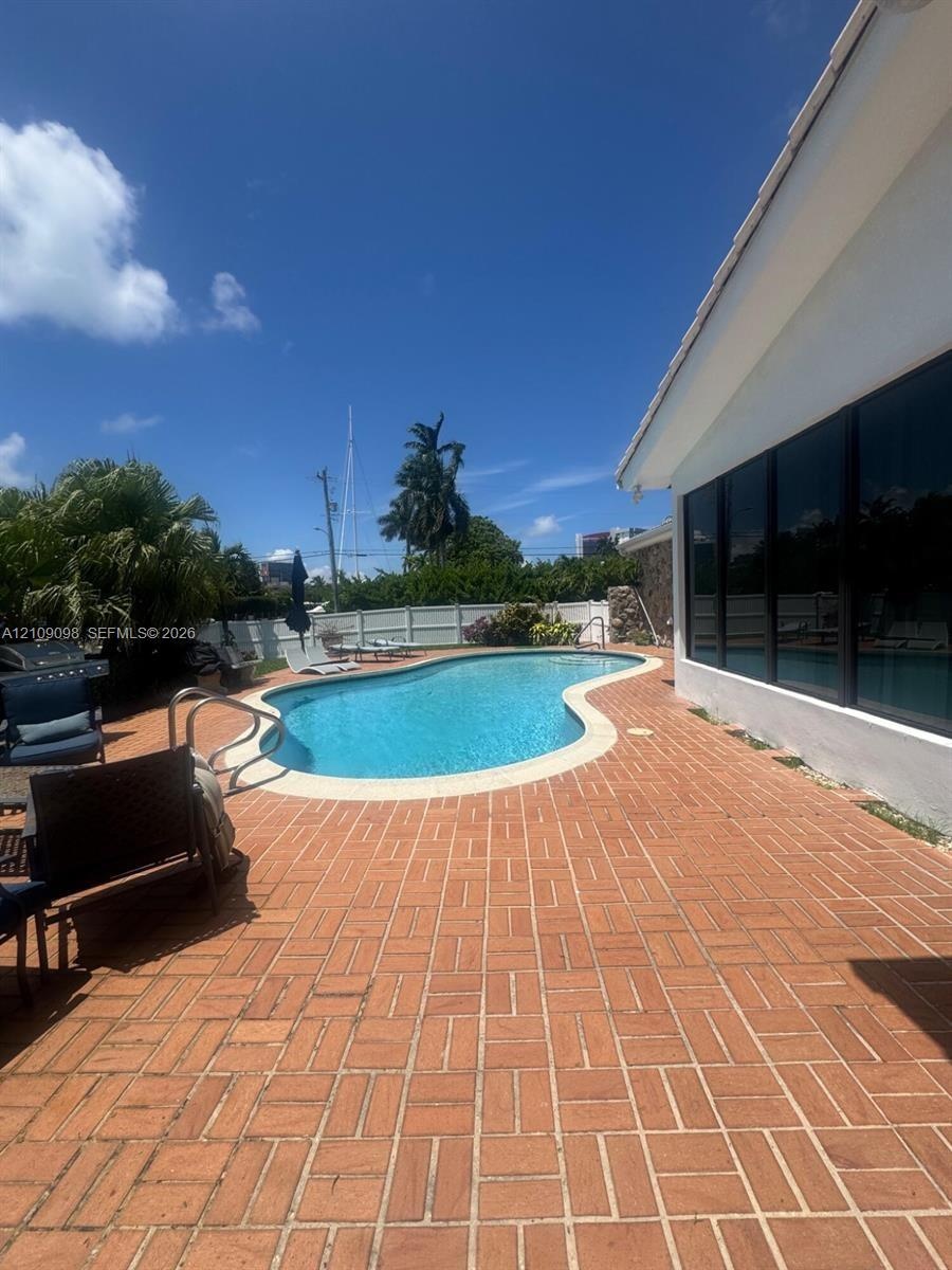 Curved in-ground swimming pool featuring a brick paver patio, full-height exterior windows, and perimeter privacy fencing