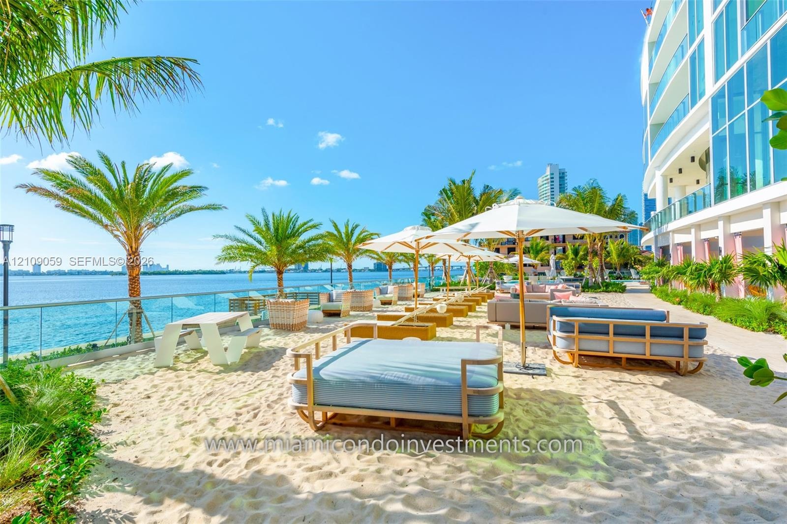 Waterfront sandy beach area featuring glass perimeter railing, mature palm trees, and structural concrete building exterior