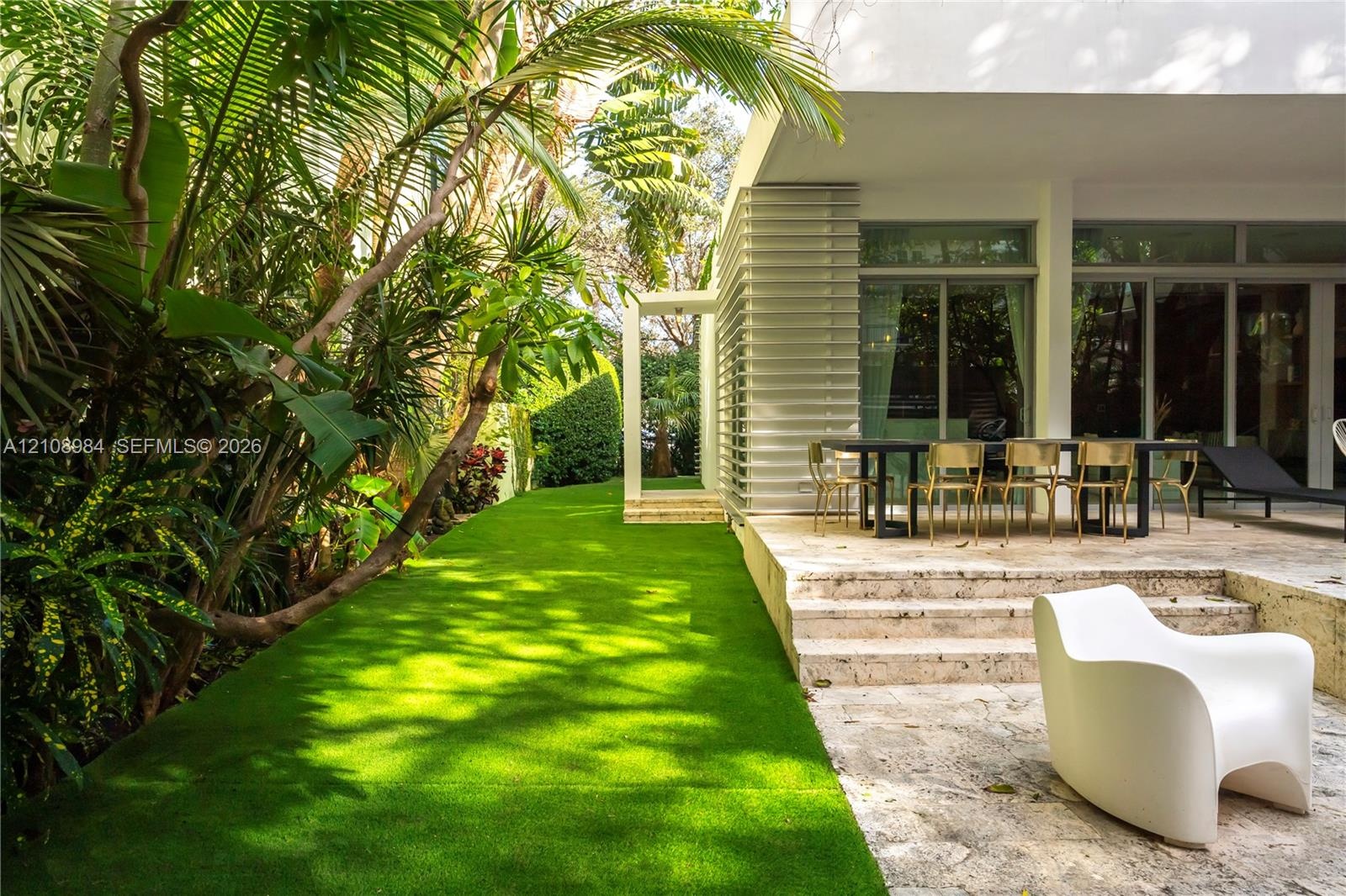Natural stone paved patio featuring a built-in step, multi-panel sliding glass doors, horizontal exterior louvers, and a bordering artificial turf law