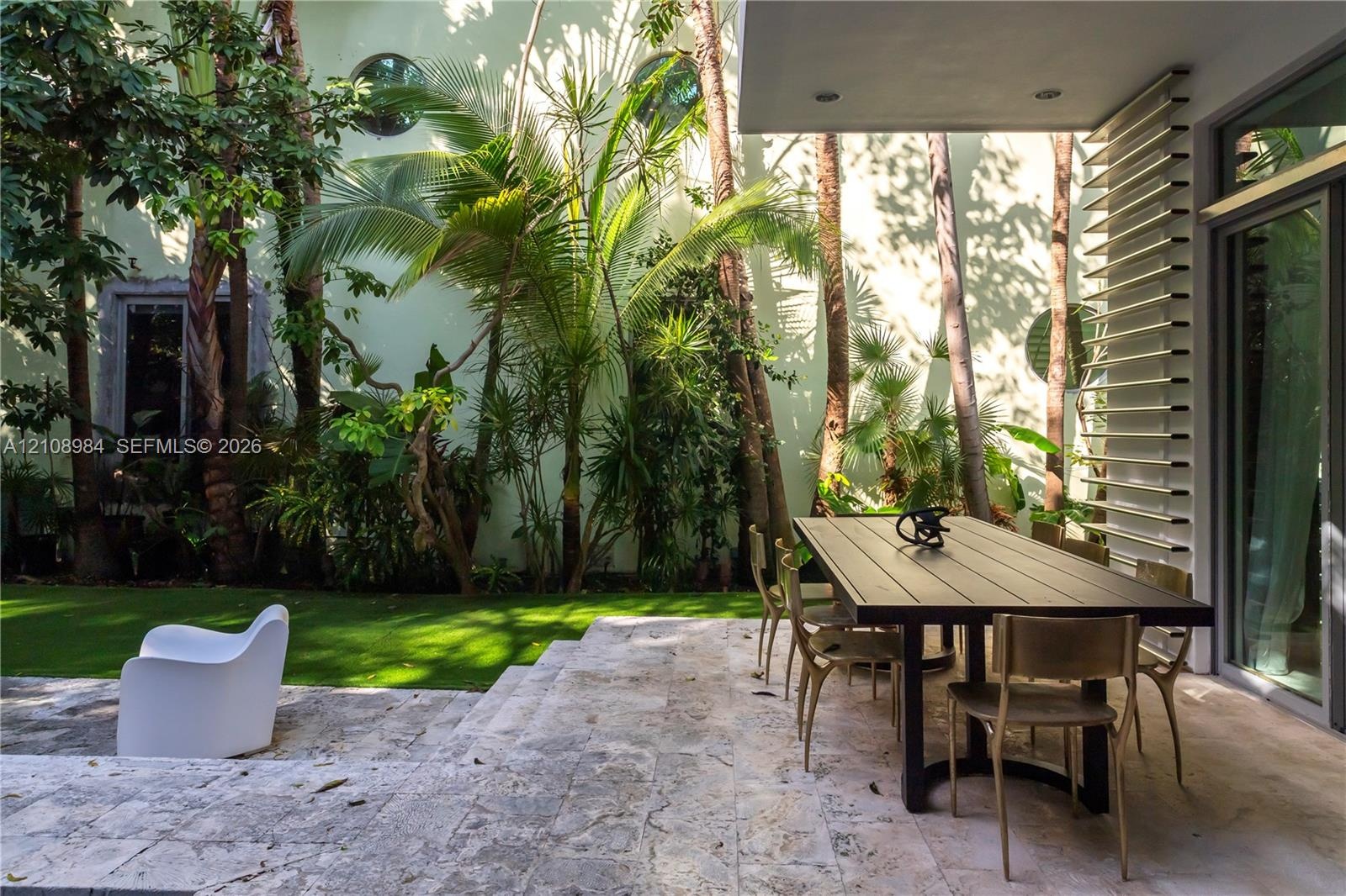 Covered patio featuring stone tile flooring, structural louvered privacy screen, synthetic turf lawn, mature palms, and circular wall accents