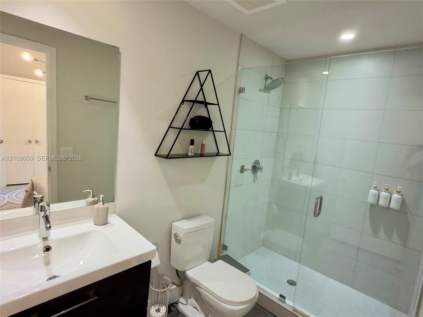 Bathroom featuring a vanity with a white countertop and undermount sink, large frameless mirror, and a walk-in shower enclosed by a glass door with li